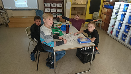 Four boys sitting around a rectangular table. Two boys are seated on milk crates. One boy is seated on a stool, while another boy is sitting in a chair.