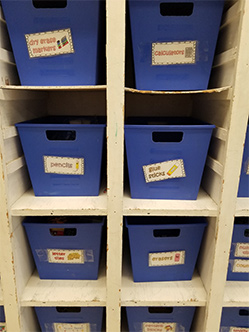 Labeled bins with cut-out handles on sectioned shelves that stand at the height of her students